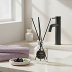 Bottle of diffuser with black sticks on a bathroom counter with towels and soap in the background