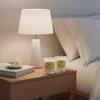 Two candles on a nightstand next to a lamp and books, with pillows in the background.