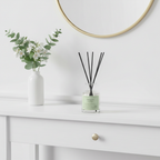 Diffuser with black sticks on a white surface, with a vase of flowers and a mirror in the background.