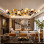 A large, ornate gold and black metal chandelier with multiple glowing globe bulbs hangs over a modern living room with a marble wall, sofa, and dark wood dining table in the foreground.