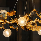 A decorative hanging chandelier with sculptural gold branches and leaves, including two spherical glass shades and several dark metallic leaf accents, shown against a dark indoor background.