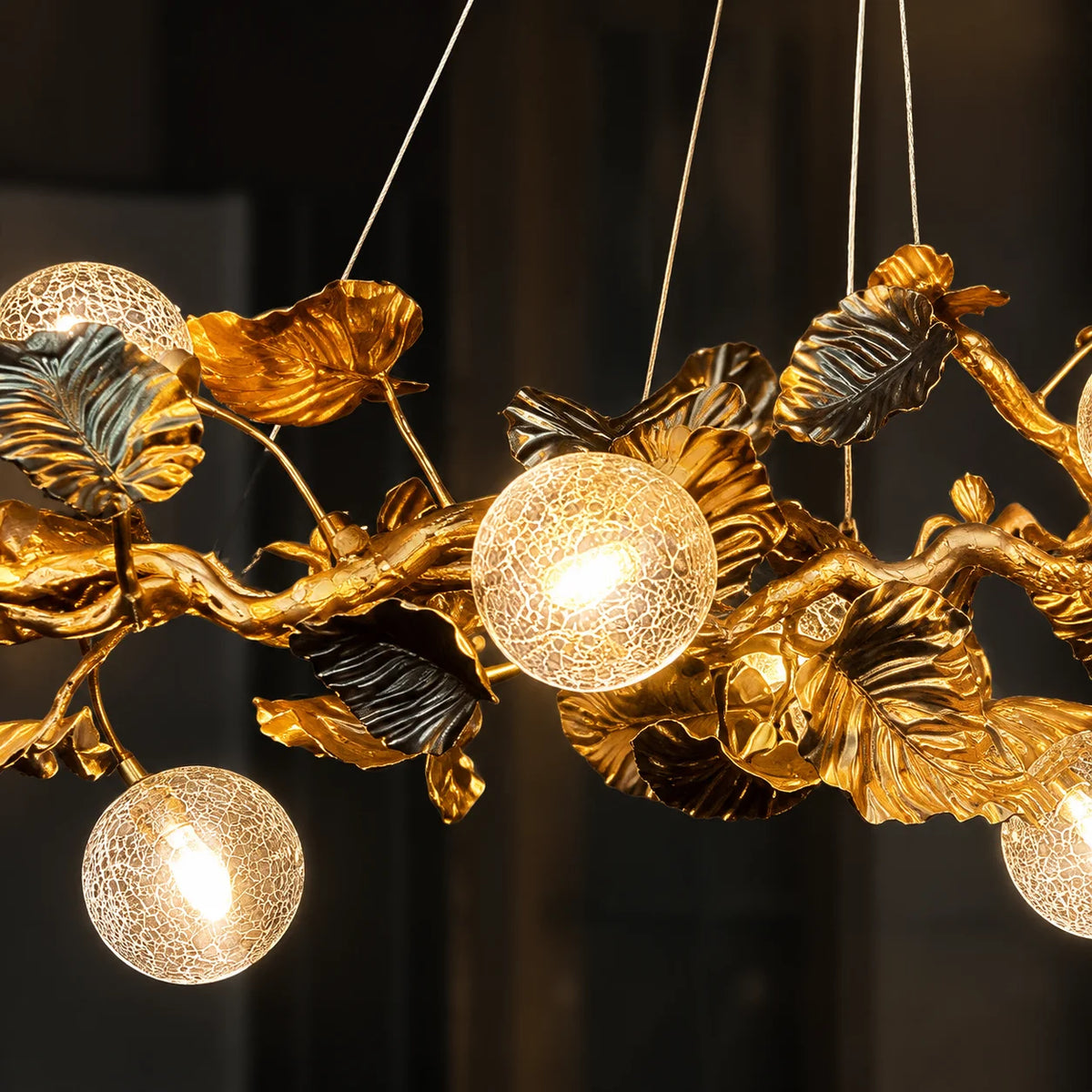 A decorative hanging chandelier with sculptural gold branches and leaves, including two spherical glass shades and several dark metallic leaf accents, shown against a dark indoor background.