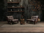 Two dark brown leather armchairs with wooden frames are arranged on a woven rug in a moody, rustic living room setting with a textured dark gray wall, floating shelves, and a small central side table.