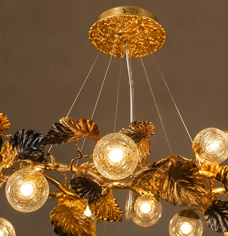 A close-up of a decorative branch-style chandelier with textured gold and dark metallic leaves and multiple glowing glass globe bulbs.