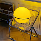 Yellow spherical object on a transparent chair in a room with wooden floor and white walls.