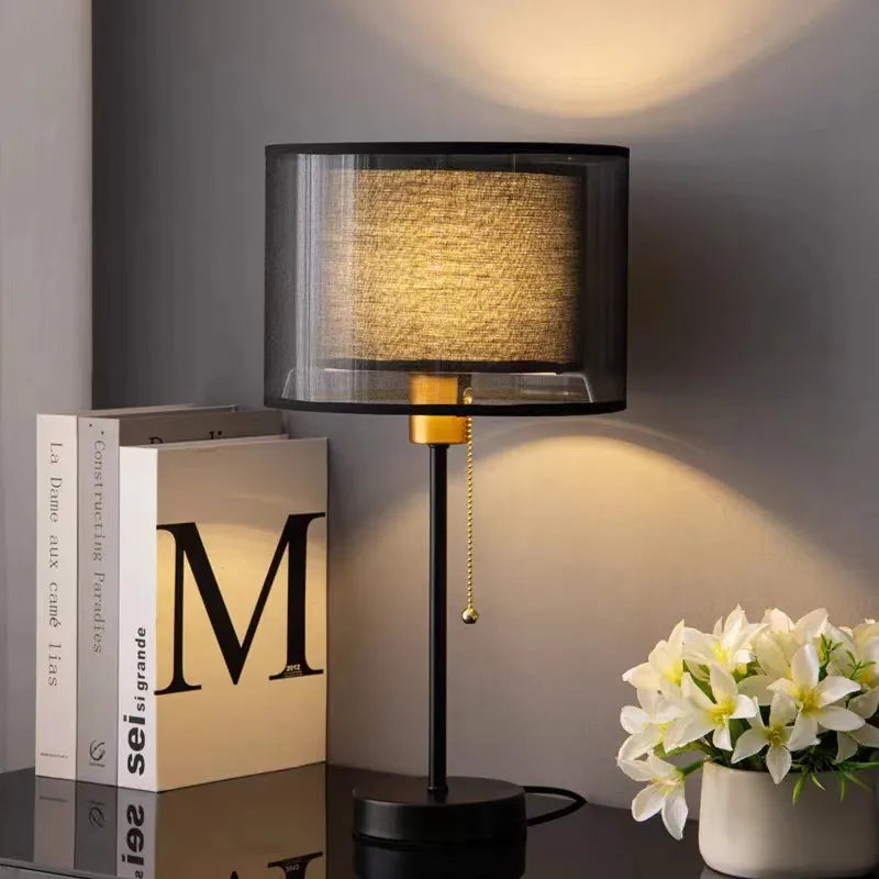 Black table lamp with a textured shade on a desk next to books and a plant.