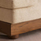 Close-up of a beige textured upholstered sofa corner with a medium brown wooden base and leg, shown on a light wood floor.