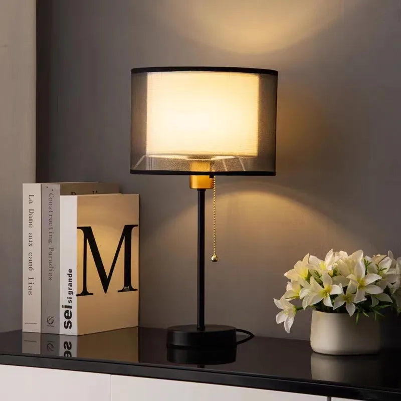 Table lamp with a black shade on a table next to books and a vase of flowers.