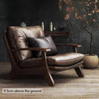 A modern lounge chair with brown leather cushions and a dark wood frame, styled in a room setting with a dark textured wall, wooden floor, and a potted tree.