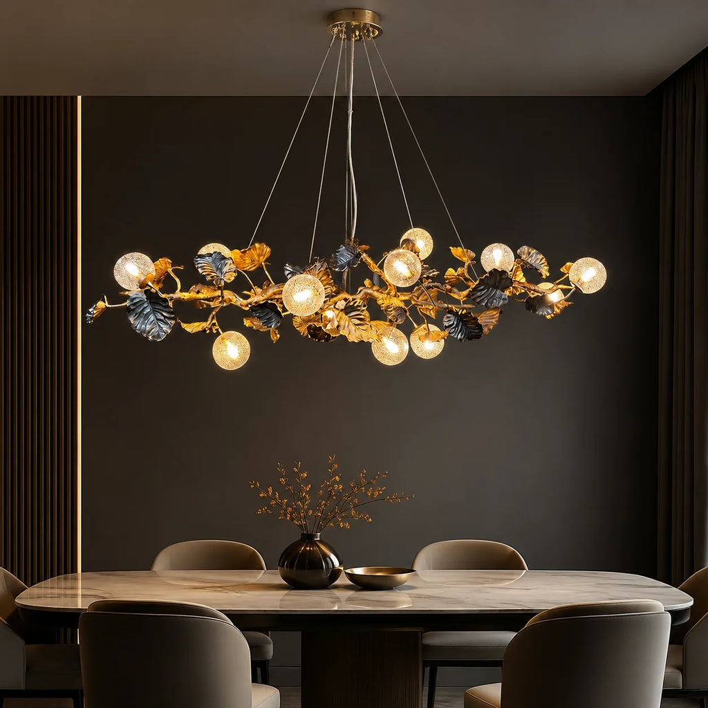A modern chandelier with a gold branch-like frame, dark blue leaf accents, and multiple glowing spherical bulbs, hanging above a dining table in a dark, elegant room.