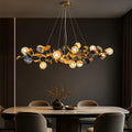A modern chandelier with a gold branch-like frame, dark blue leaf accents, and multiple glowing spherical bulbs, hanging above a dining table in a dark, elegant room.
