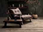 A modern lounge chair with tufted taupe-gray cushions and curved dark wood arms, styled in a rustic room with a matching wooden coffee table, textured gray wall, and a potted tree.