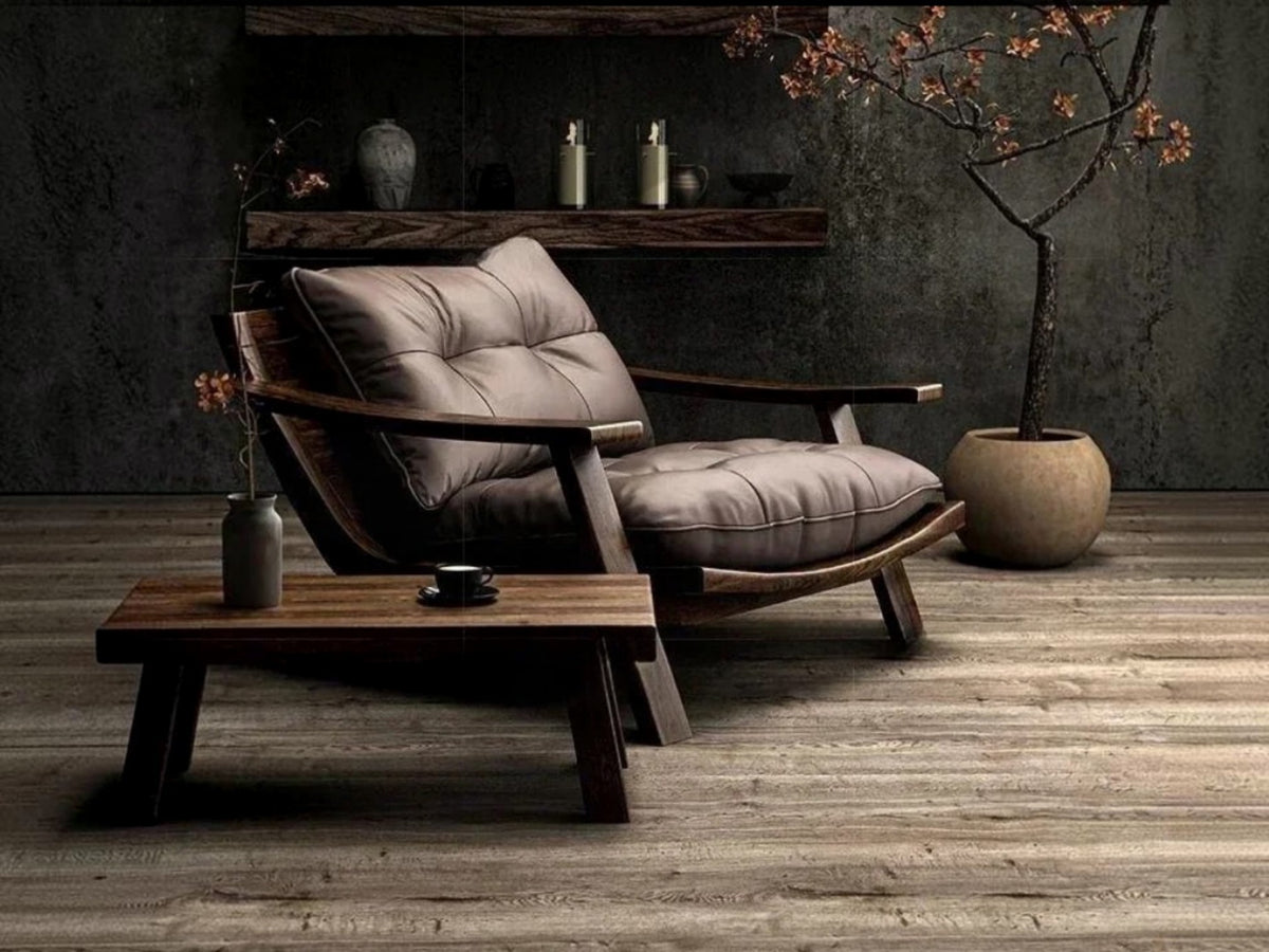 A modern lounge chair with tufted taupe-gray cushions and curved dark wood arms, styled in a rustic room with a matching wooden coffee table, textured gray wall, and a potted tree.