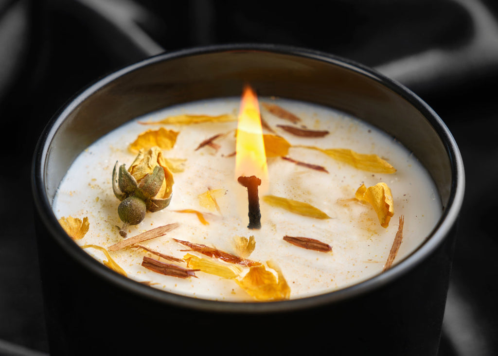 Candle in a black mug with a white candle wick, surrounded by dried herbs and flowers on a dark background.
