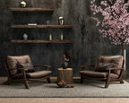 A styled interior scene with two brown leather armchairs facing a small round wooden side table, set on a textured rug against a dark textured wall with floating shelves and pink flowering branches on the right.