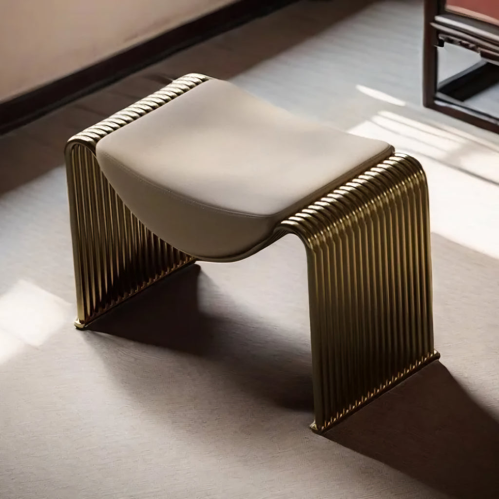 A modern sculptural bench with a curved cream upholstered seat and a ribbed metallic gold frame, shown in a sunlit interior setting.