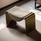 A modern sculptural bench with a curved cream upholstered seat and a ribbed metallic gold frame, shown in a sunlit interior setting.