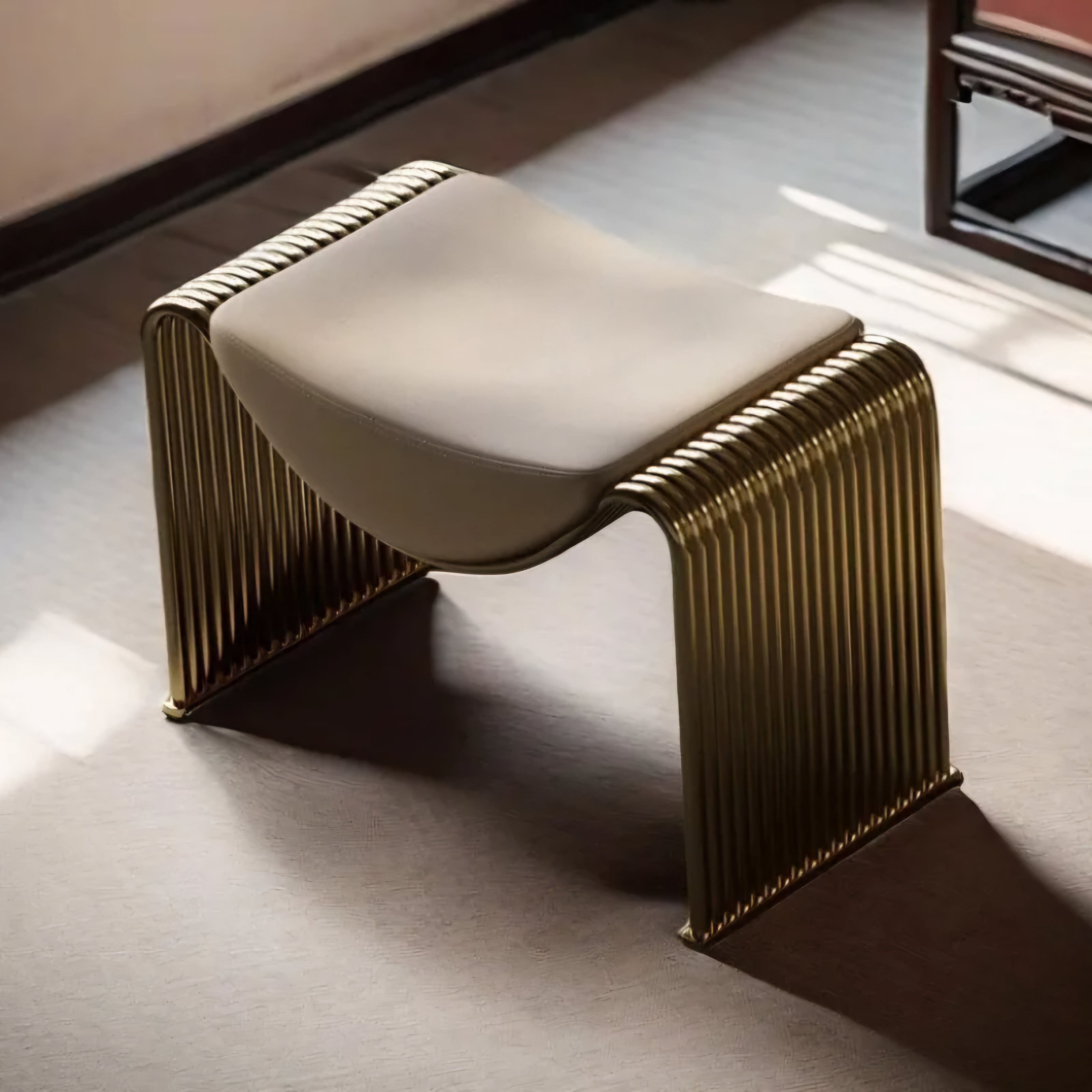 A modern sculptural bench with a curved cream upholstered seat and a ribbed metallic gold frame, shown in a sunlit interior setting.