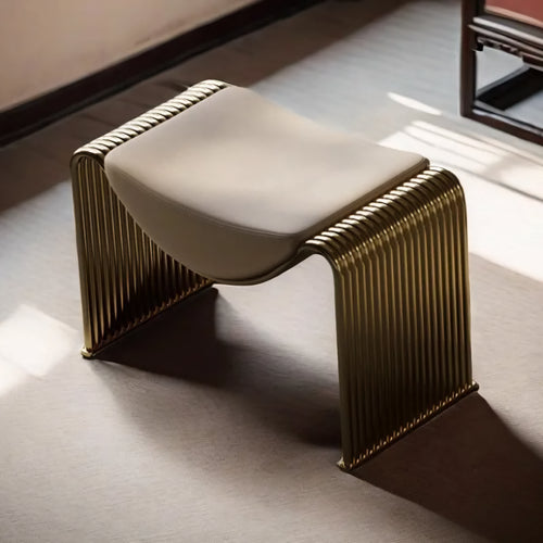 A modern sculptural bench with a curved cream upholstered seat and a ribbed metallic gold frame, shown in a sunlit interior setting.