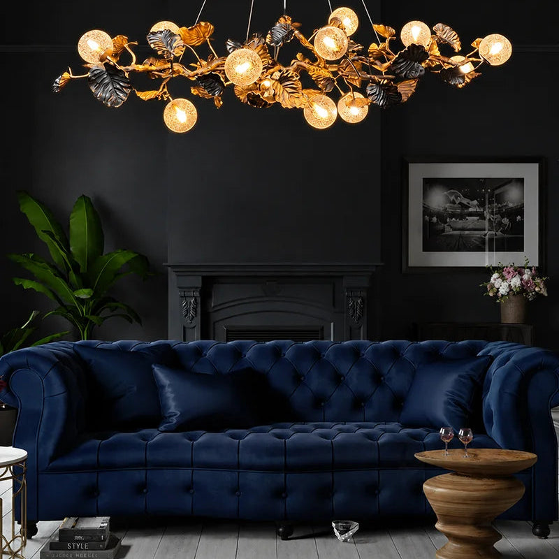 A luxurious gold and black leaf-shaped chandelier with glowing globe bulbs hangs in a dark, elegant living room above a deep navy blue tufted sofa.