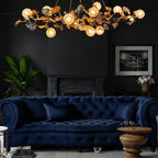 A luxurious gold and black leaf-shaped chandelier with glowing globe bulbs hangs in a dark, elegant living room above a deep navy blue tufted sofa.