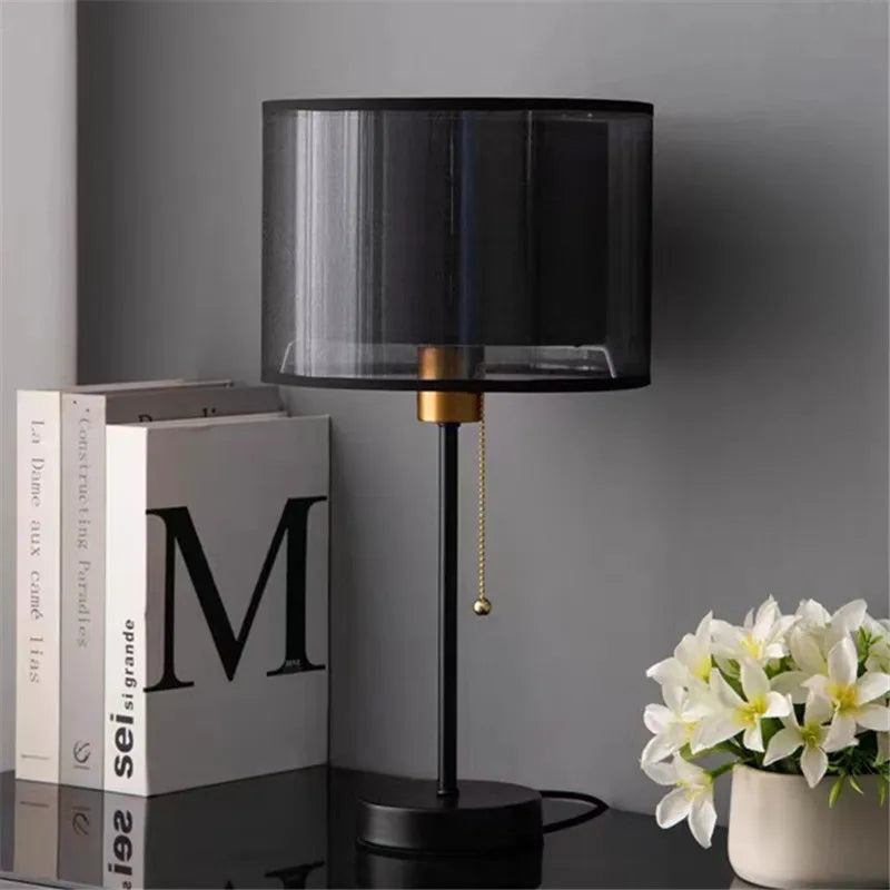 Black table lamp with a gold accent on a dark surface next to books and a vase of flowers.