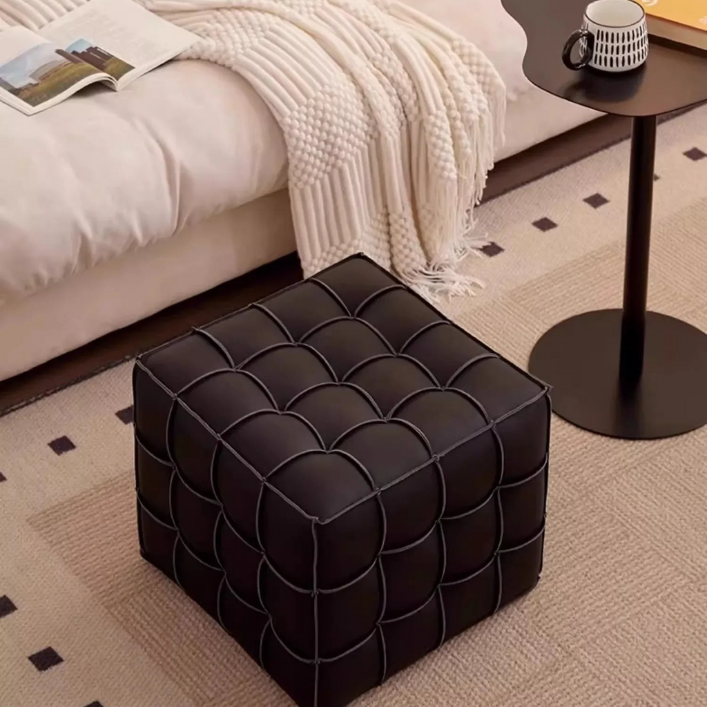 Black tufted ottoman in a bedroom setting with a bed and side table.