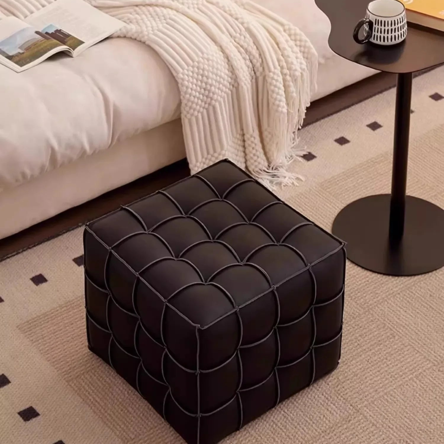 Black tufted ottoman in a bedroom setting with a bed and side table.