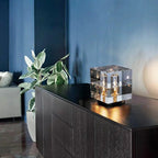 Modern interior with a black cabinet, clear crystal cube lamp, and green plant against a blue wall.