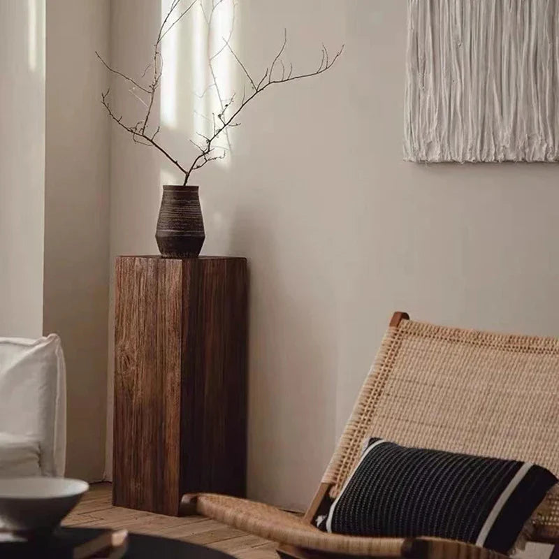 Wicker chair with a black and white pillow next to a wooden pedestal with a vase and branches, against a neutral wall.