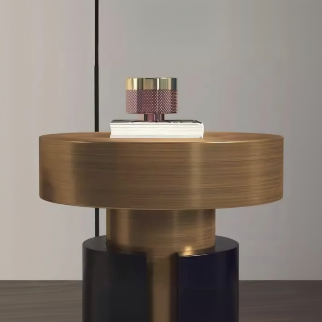 A modern round table lamp with a brushed brass-finished cylindrical body, a dark navy base, and a small white book on top of the lamp base, set against a neutral gray background with promotional text at the bottom.