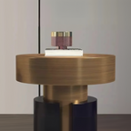 A modern round table lamp with a brushed brass-finished cylindrical body, a dark navy base, and a small white book on top of the lamp base, set against a neutral gray background with promotional text at the bottom.