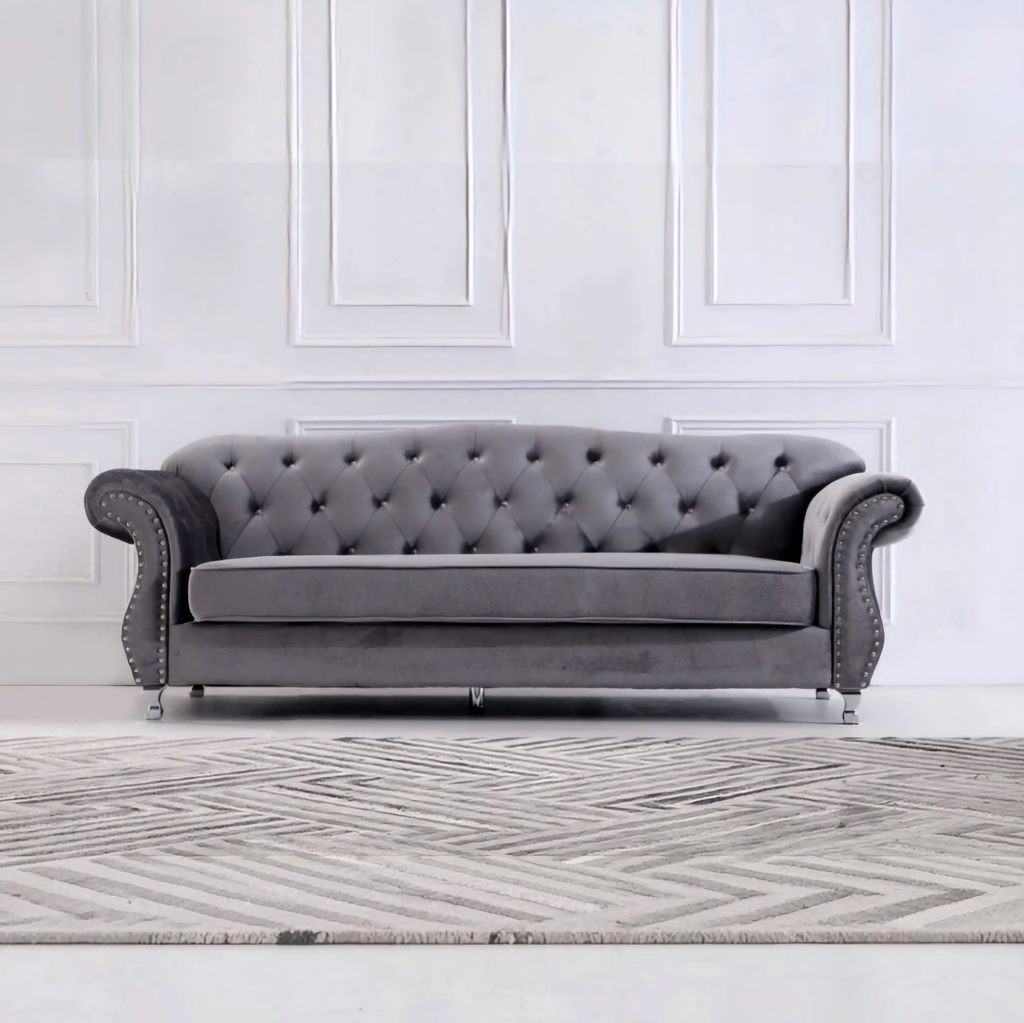 A classic gray velvet Chesterfield loveseat sofa with tufted backrest, rolled arms, and silver nailhead trim, positioned in front of a white paneled wall and a gray patterned rug.
