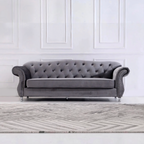 A classic gray velvet Chesterfield loveseat sofa with tufted backrest, rolled arms, and silver nailhead trim, positioned in front of a white paneled wall and a gray patterned rug.