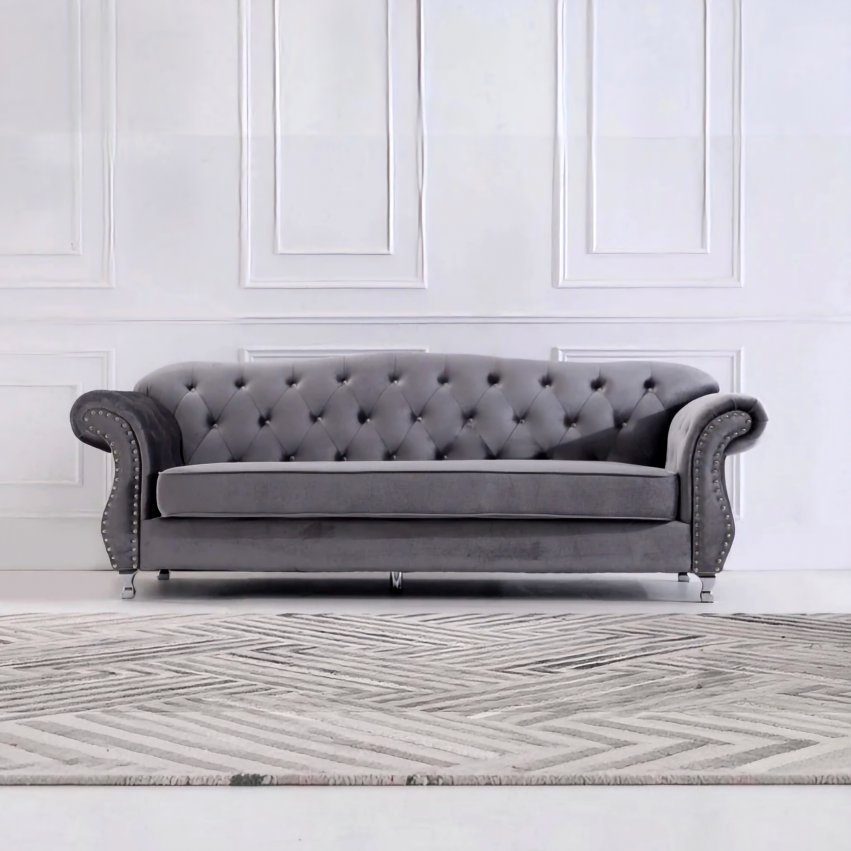 A classic gray velvet Chesterfield loveseat sofa with tufted backrest, rolled arms, and silver nailhead trim, positioned in front of a white paneled wall and a gray patterned rug.