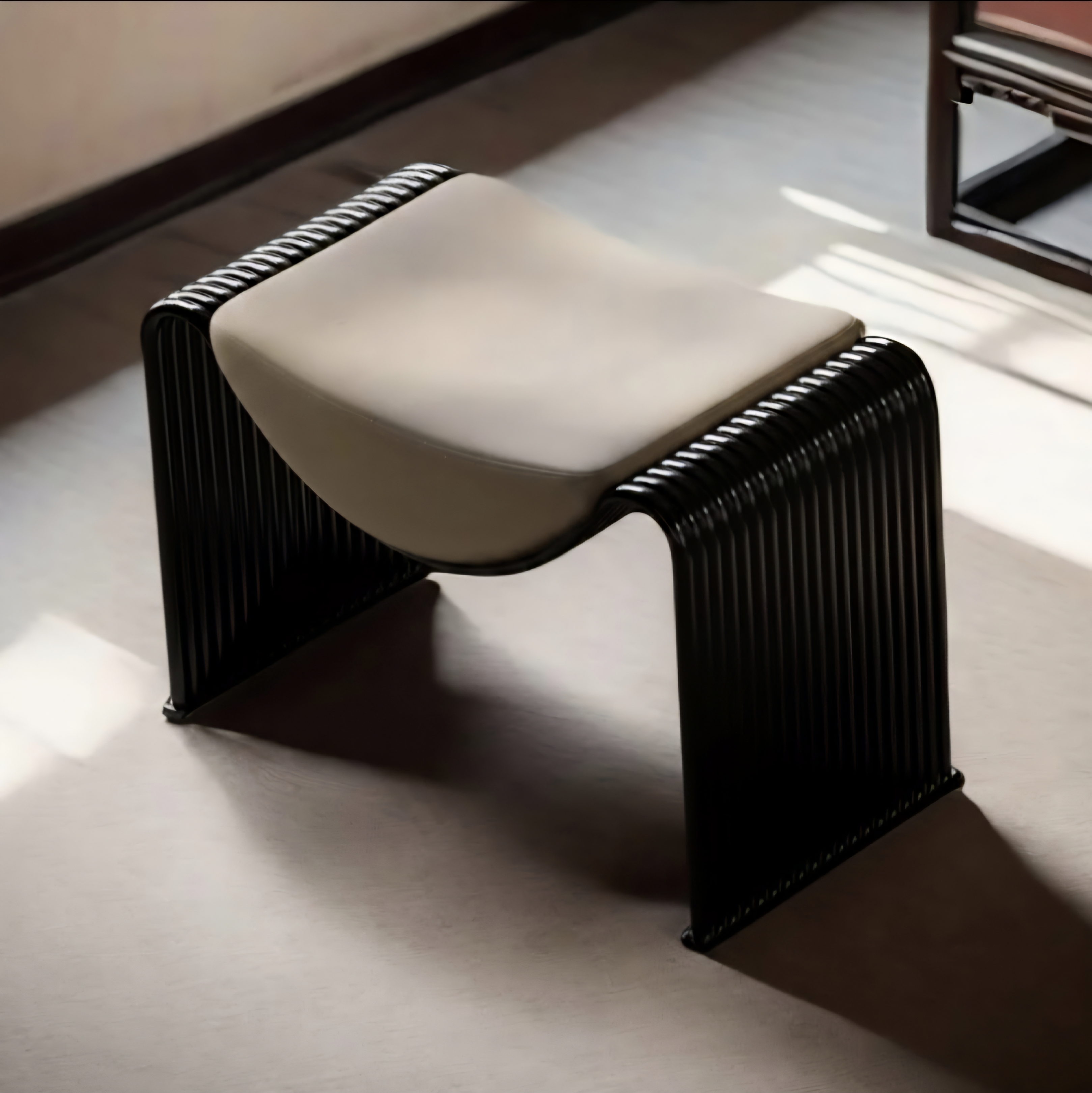 A modern sculptural bench or stool with a curved cream cushion and a black ribbed metal frame, photographed in a softly lit interior setting.