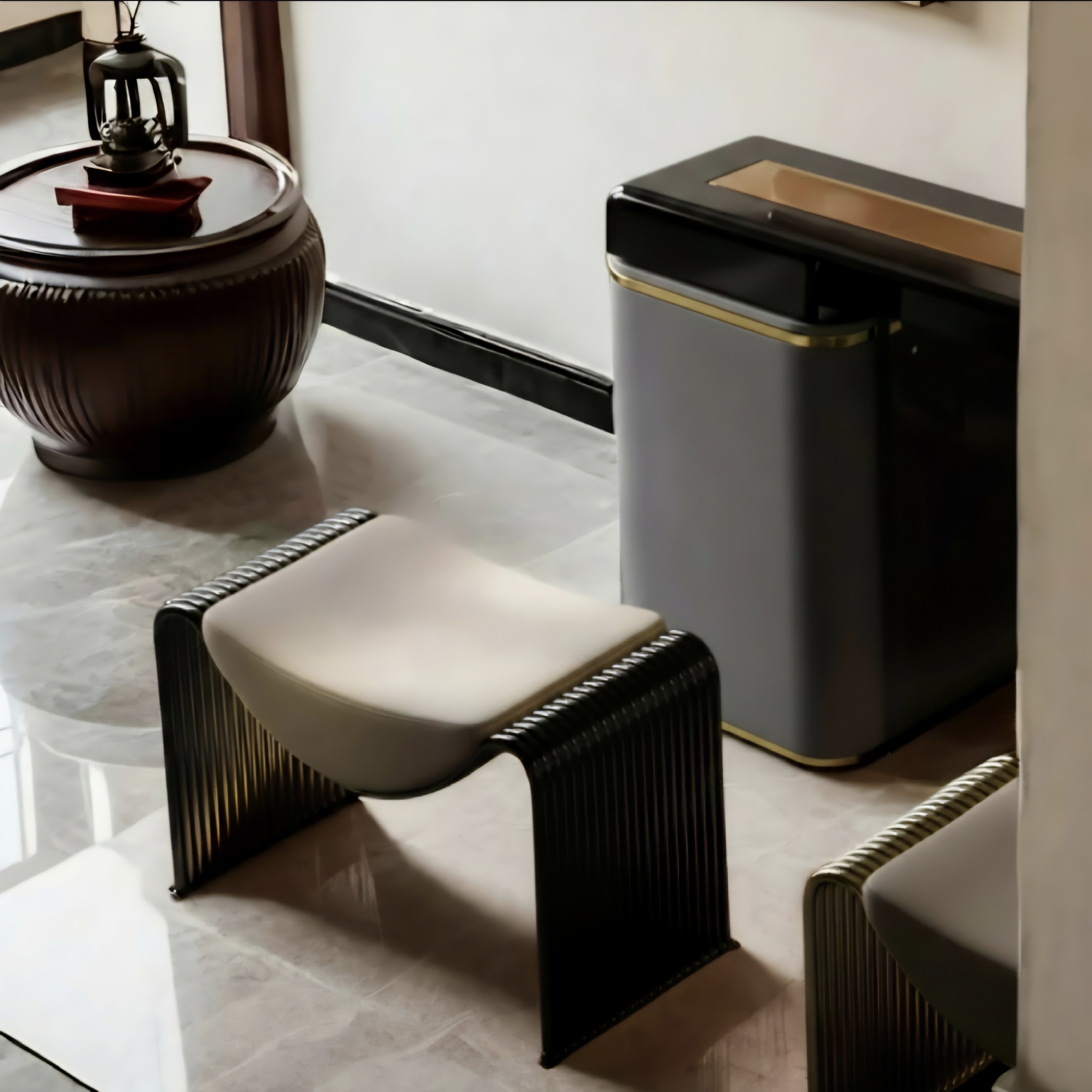 A modern black ribbed metal stool with a cream cushion sits in a minimalist interior next to a dark gray trash can with gold trim.