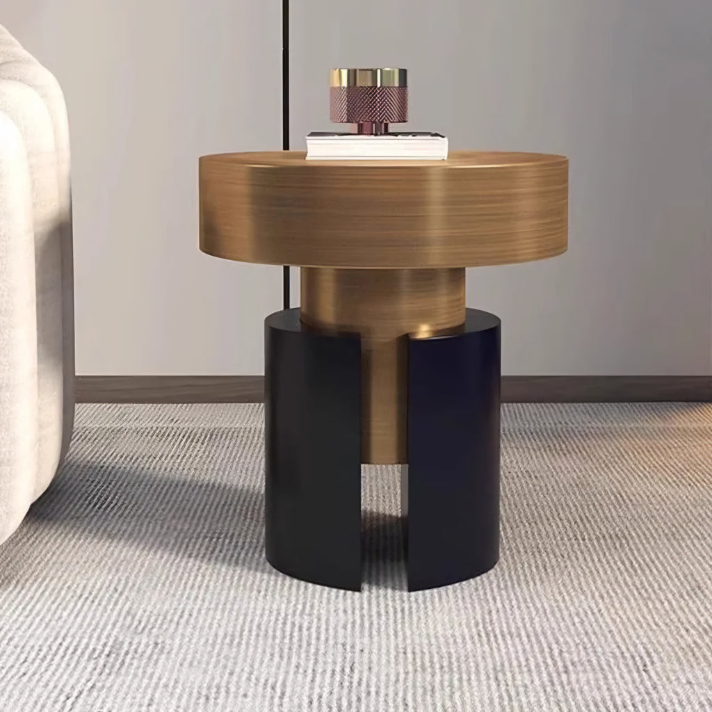 A modern cylindrical side table with a brushed gold top and base, and a split black pedestal base, shown in a styled interior setting next to a sofa.