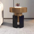 A modern cylindrical side table with a brushed gold top and base, and a split black pedestal base, shown in a styled interior setting next to a sofa.
