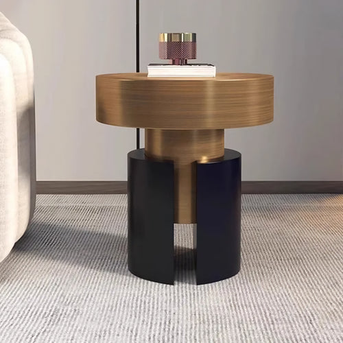 A modern cylindrical side table with a brushed gold top and base, and a split black pedestal base, shown in a styled interior setting next to a sofa.