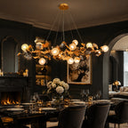 A large, ornate gold chandelier with glowing spherical bulbs and leaf-like details hangs over a dining table in a dark, elegant dining room with a fireplace and arched doorway.
