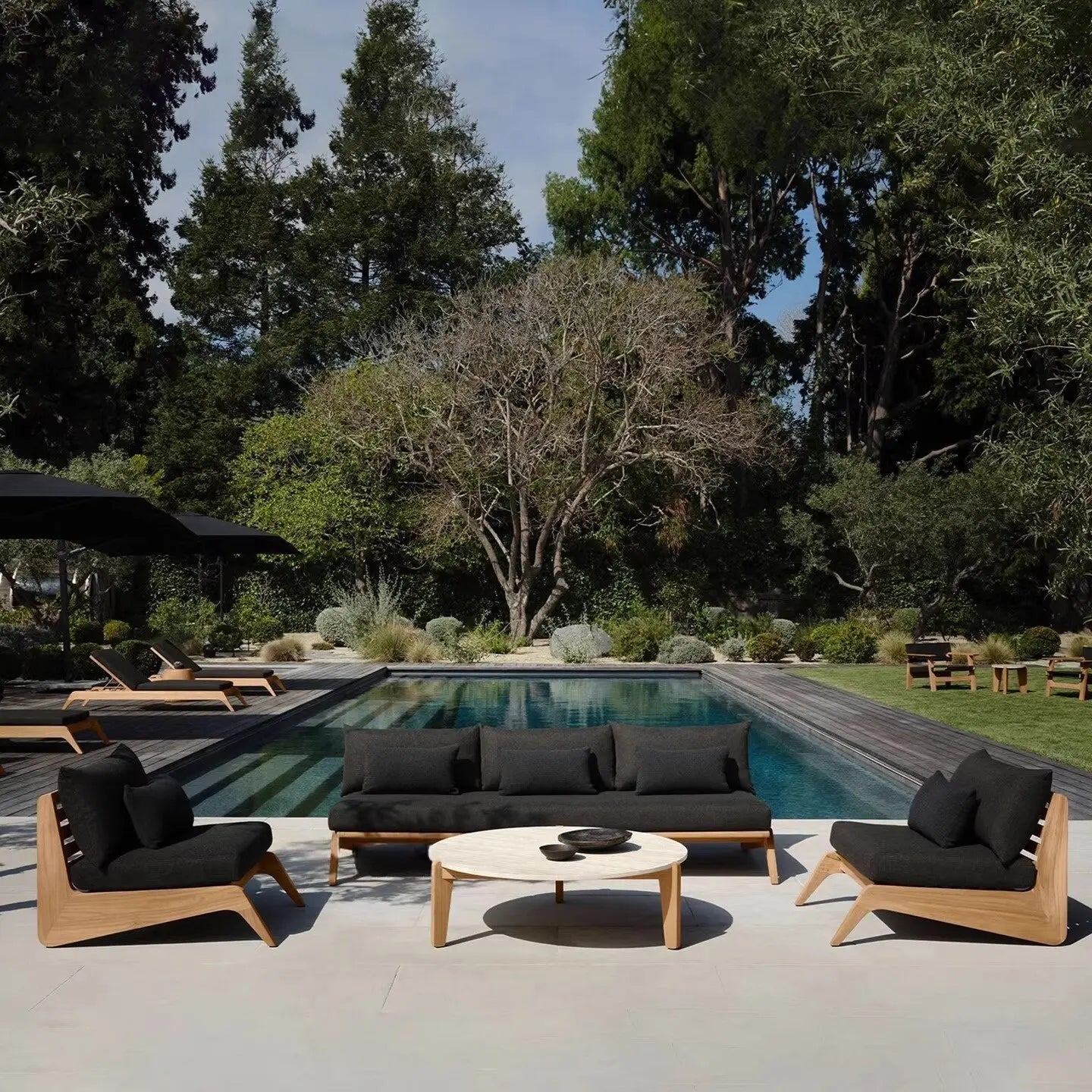 Outdoor patio set with black cushions by a pool surrounded by trees