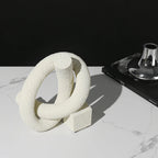 White textured ring sculpture on a marble surface with a black book and metallic object in the background.