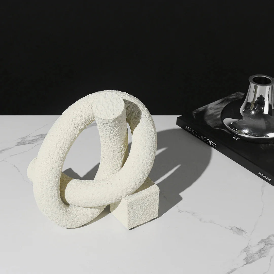 White textured ring sculpture on a marble surface with a black book and metallic object in the background.