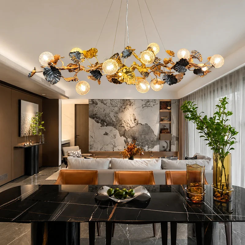A modern dining room featuring a large, decorative leaf-shaped chandelier with glowing globe bulbs hanging over a black marble dining table. The background shows a contemporary living room with a marble wall art piece, white sofa, and warm lighting.