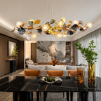 A modern dining room featuring a large, decorative leaf-shaped chandelier with glowing globe bulbs hanging over a black marble dining table. The background shows a contemporary living room with a marble wall art piece, white sofa, and warm lighting.
