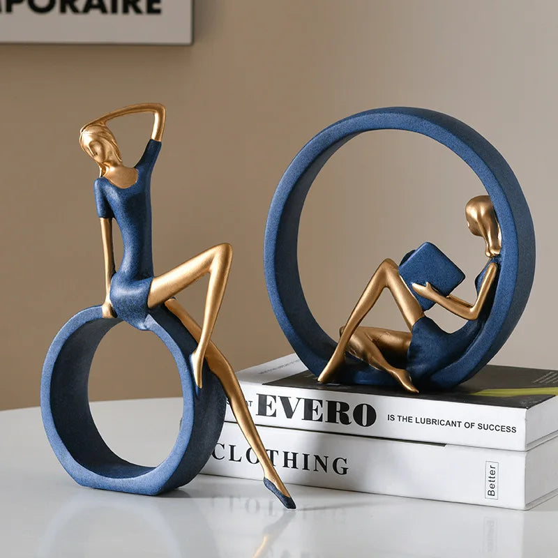 Two decorative figurines shaped like seated women in blue dresses and gold metallic limbs, displayed on a white tabletop with stacked books. One figurine sits on a large blue ring while the other is inside a circular blue frame reading a book.