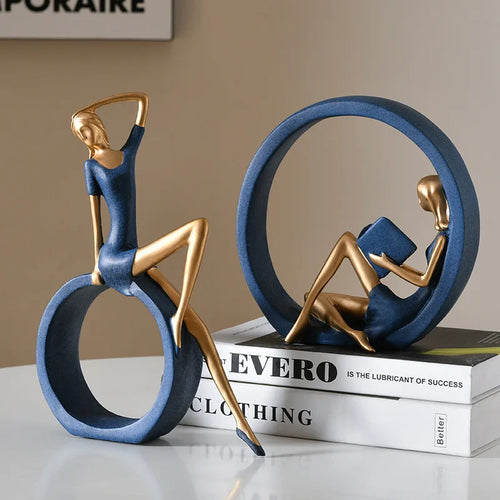 Two decorative figurines shaped like seated women in blue dresses and gold metallic limbs, displayed on a white tabletop with stacked books. One figurine sits on a large blue ring while the other is inside a circular blue frame reading a book.