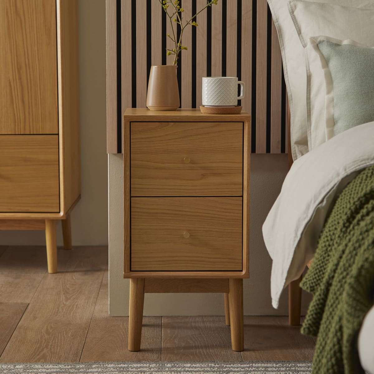 Wooden nightstand with two drawers, a vase, and a mug in a bedroom setting.