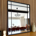 Large wall mirror with black frame reflecting a dining room with wooden chairs and a chandelier.